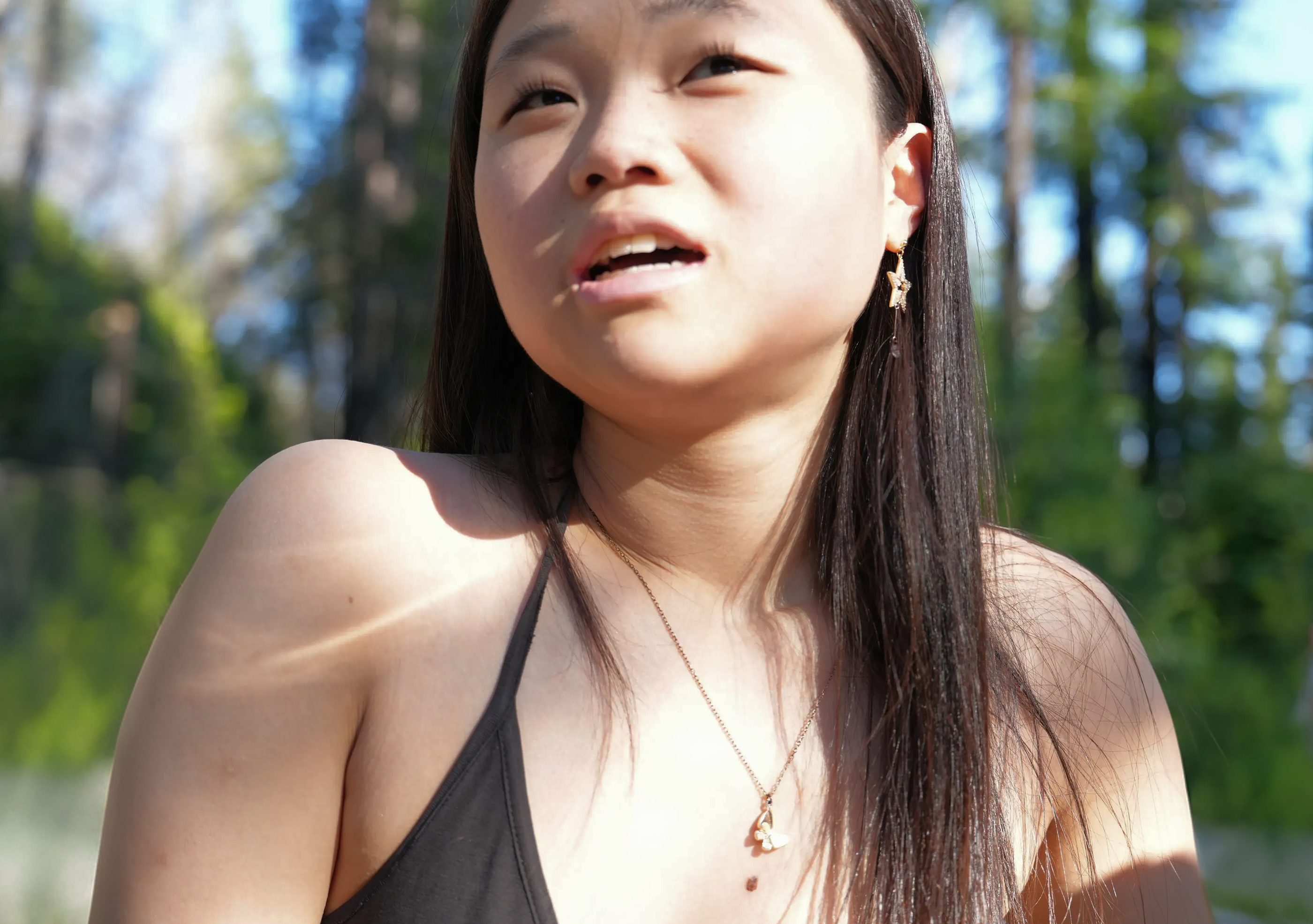 A woman looking up in dappled sunlight among tall trees.