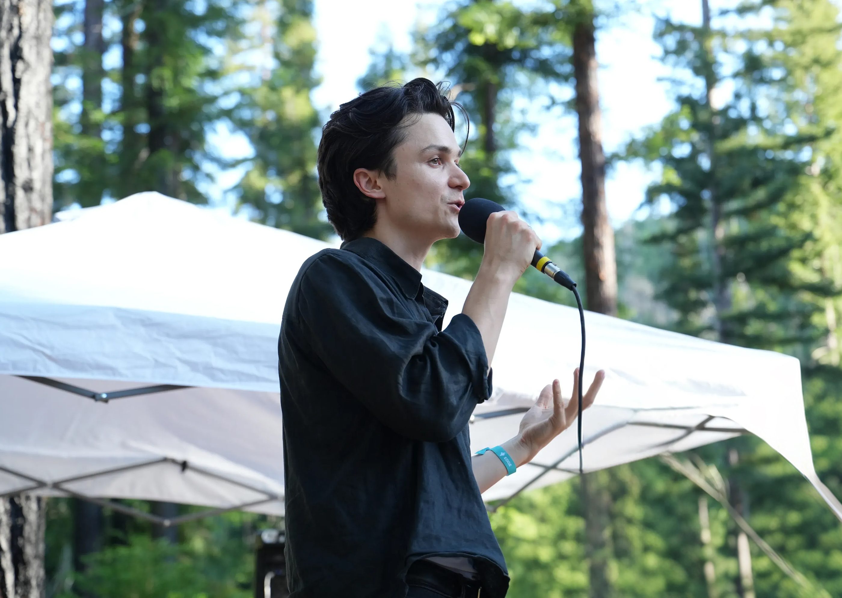 A person speaking into a microphone under a white canopy, framed by redwoods.