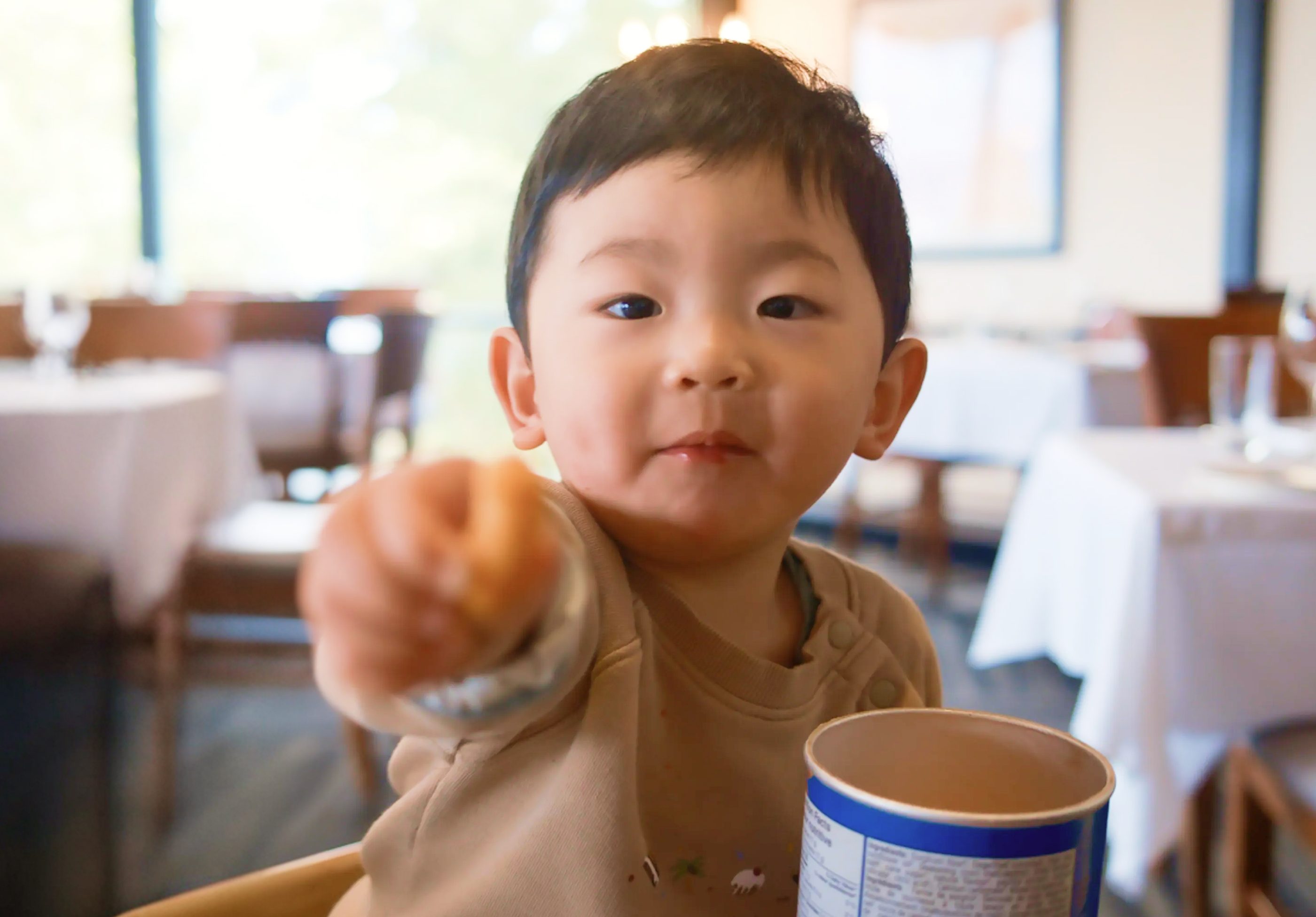 A toddler in a sunlit restaurant pointing at the camera.