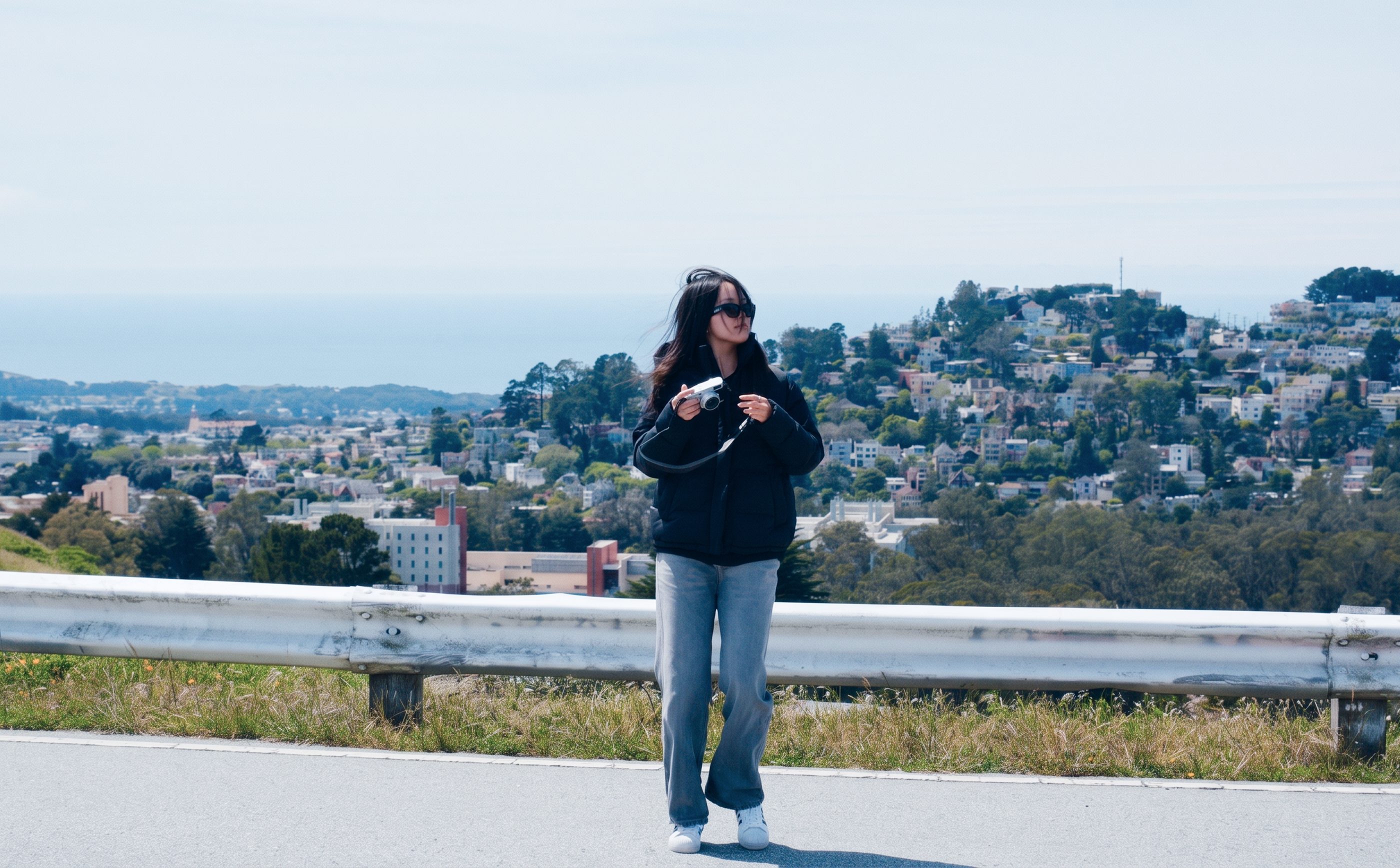Amy walking along a road with the hills of San Francisco behind her, holding a camera.